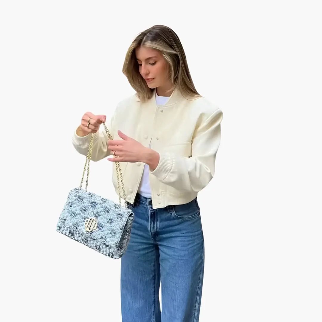 Cream cropped bomber jacket with button closure and ribbed hem; blue patterned handbag on chain strap.